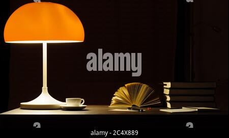 A table lamp with an orange lampshade lights up the books on the desk Stock Photo
