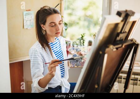 Thoughtful creative girl in white shirt and striped T-shirt holding paint brush in hand while dreamily drawing on easel at home Stock Photo