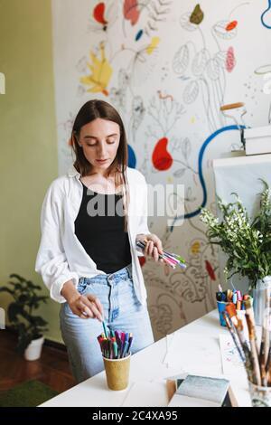 Pretty girl near desk dreamily holding colored felt-tip pens in hand with patterns canvas on background at home Stock Photo