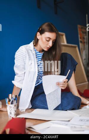 Thoughtful girl in white shirt and striped T-shirt sitting on floor and holding piece of paper in hand with canvases on background at home Stock Photo