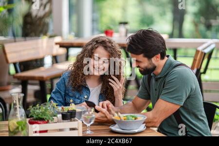 Happy couple sitting outdoors on terrace restaurant, using smartphone. Stock Photo
