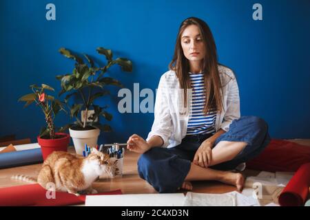 Pensive girl sitting on floor with drawings frightenedly looking at cat near while spending time at home Stock Photo