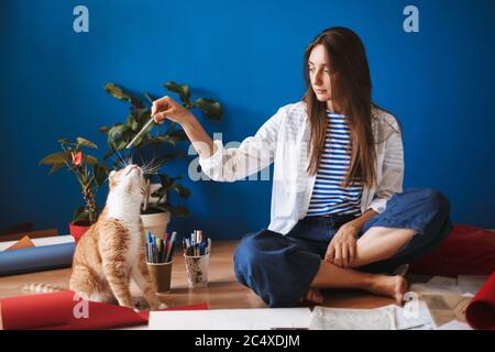 Beautiful girl sitting on floor holding pencil in hand dreamily playing with cat while spending time at cozy home Stock Photo