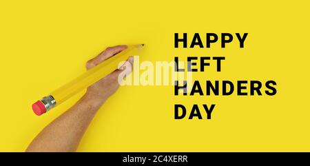 Big pencil in male left hand on yellow background with text Happy Left Handers Day. Conceptual banner for celebration of international left-handed day Stock Photo