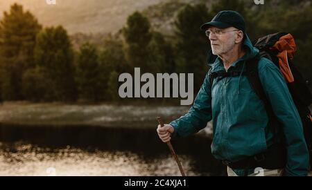 Senior man walking by a river on mountain trail. Fit mature man carrying a backpack hiking in nature. Stock Photo