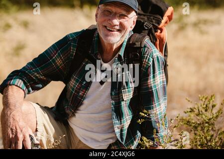 Portrait of a senior man carrying a backpack looking at camera and smiling. Retired male hiker taking a break, sitting on ground. Stock Photo