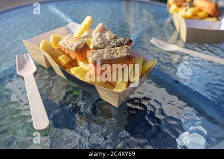 fish and chips with fried smoked salmon and french fries in a cardboard box and a wooden fork on a glass table, fast food in an outdoor street restaur Stock Photo