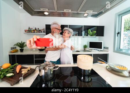 Happy senior couple taking selfie while cooking together at home - Mature people having fun preparing the lunch - Joyful elderly lifestyle and food nu Stock Photo