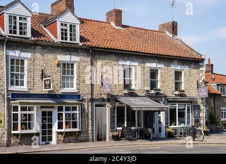 Village street with a row of stone-made buildings with shops.  A bicycle rests against a bollard and containers with flowers stand on the pavement. Stock Photo