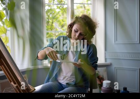 Adult woman squeezing paint from bottle on palette while working in cozy studio Stock Photo