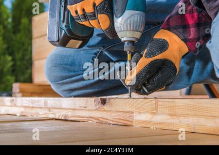 Male Carpenter Using Cordless Drill To Drive Long Screw Through Wooden Planks. Stock Photo