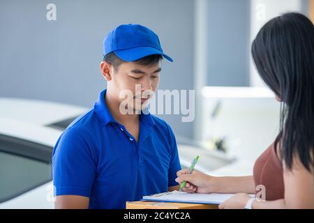 Asian delivery servicemen wearing a blue uniform with a bluecap and  handling cardboard boxes to give to the female customer in front of the house. On Stock Photo