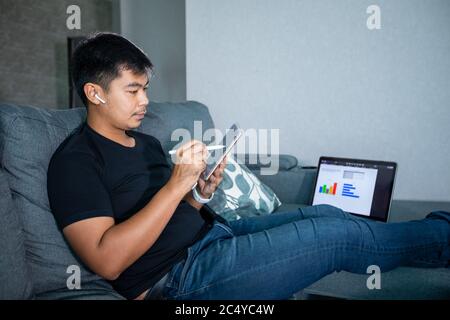 Asian businessman using wireless earphones and using computer notebook and tablet working from home.He is prepare graphs and information for online me Stock Photo