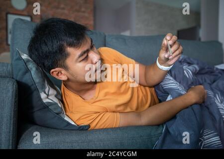 Asian man is ill and coughs on the sofa at home and he uses a thermometer to check the temperature during the coronavirus epidemic. Stock Photo