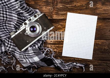 Old retro camera on vintage brown wooden board with black and white checkered scarf and piece of squared paper for notes. abstract background. Top vie Stock Photo