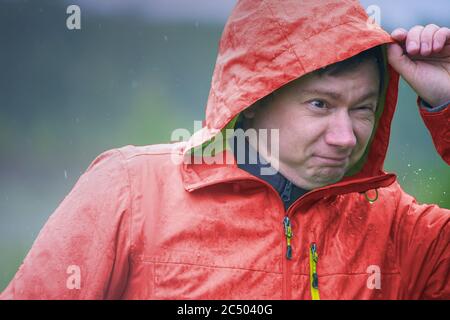 A young guy in a raincoat with a hood stands in the rain. Raindrops fall on your face and jacket. Stock Photo