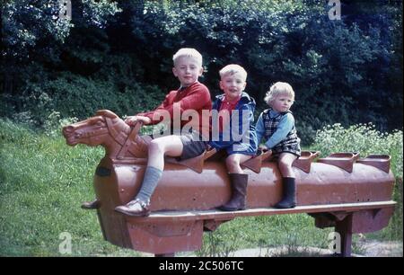 1967, three young children playing or riding on a brown coloured metal five seater rocking horse outside in a park, Surrey, England, UK. This type of traditional, possibly victorian, play equipment was to be found in playgrounds and public parks across Britain in this era and young children had enormous fun playing on them. Stock Photo