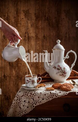 Hand pouring milk into coffee served on vintage tableware with homemade cookies on rustic table with a wooden background. Stock Photo