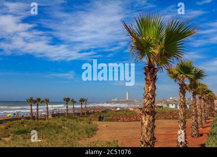 Palm trees along the Atlantic Ocean shore and unidentifiable people walking on the beach with the lighthouse in the background in Casablanca, Morocco Stock Photo