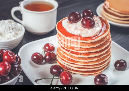 Morning breakfast of homemade pancakes with curd or cottage cheese, juicy ripe cherries, flowing maple syrup and a cup of tea on a wooden rustic table Stock Photo