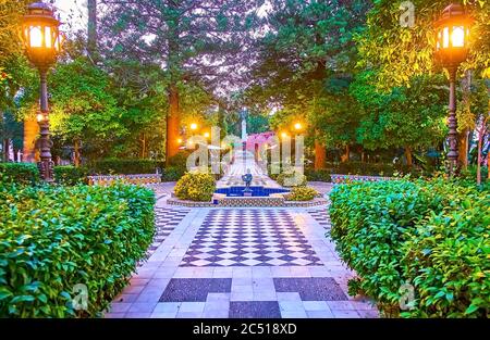 Evening in topiary Alameda Apodaca and Marques de Comillas garden with chessboard pattern floor, spread trees, trimmed bushes, cast iron retro lantern Stock Photo