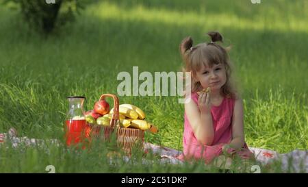 Weekend at picnic. Toddler caucasian girl on green grass meadow with basket full of fruits. Eating pancakes. Female child kid in pink dress on nature sit on white blanket having fun. Sunny summer Stock Photo