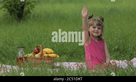 Weekend at picnic. Lovely caucasian girl on green grass meadow with basket full of fruits. Waving her hands. Female child kid in pink dress on nature sit on white blanket having fun. Sunny summer Stock Photo