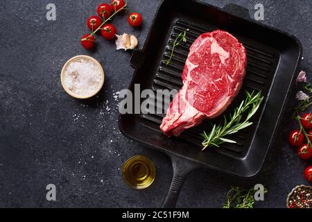 Top view Black Angus prime beef rib eye steak on cast iron grill pan with fresh rosemary, cherry tomatoes, olive oil and spices. Creative layout with