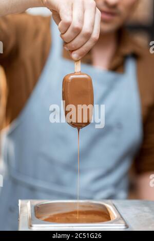 Seller in apron dips ice cream into the hot melted chocolate, making ice cream on a stick with toppings, close-up Stock Photo