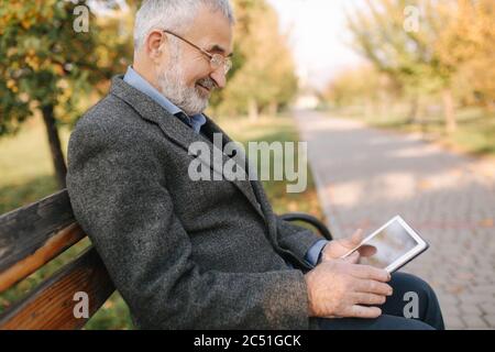Mock up of senior man using tablet outside. Back view of elderly man sitting on the bench and using tablet Stock Photo