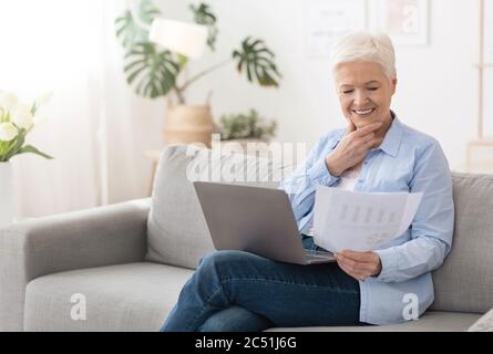 Senior woman using laptop and reading documents while working remotely at home Stock Photo
