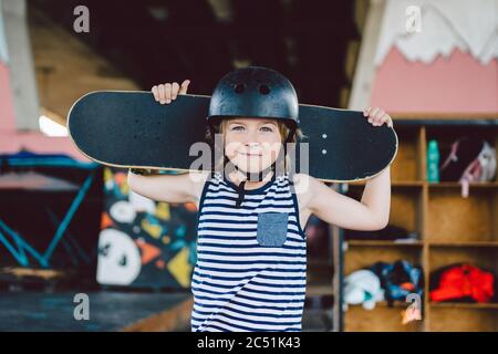Portrait of handsome caucasian boy athlete skateboarder in protective helmet with skateboard in hands looking at camera on background of skate park. A Stock Photo