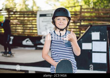 Portrait of handsome caucasian boy athlete skateboarder in protective helmet with skateboard in hands looking at camera on background of skate park. A Stock Photo