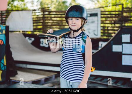 Portrait of handsome caucasian boy athlete skateboarder in protective helmet with skateboard in hands looking at camera on background of skate park. A Stock Photo