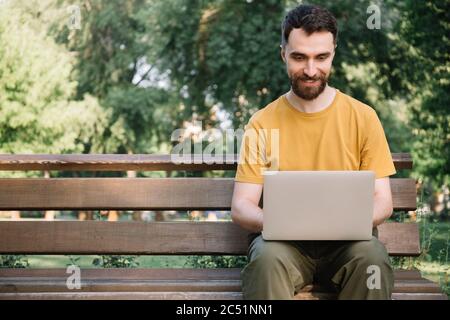 Bearded man using laptop computer, shopping online, sitting on bench. Successful freelancer working in park, typing on keyboard Stock Photo