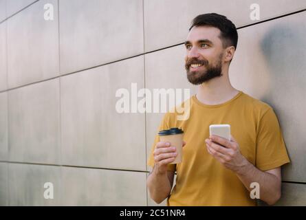 Young bearded holding smartphone and cup of coffee, shopping online. Smiling hipster using mobile phone, waiting for taxi outdoors Stock Photo