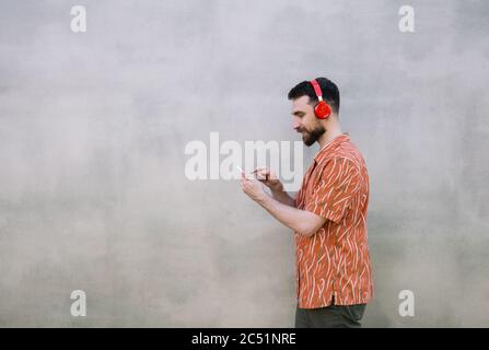 Young bearded man wearing red headphones, listening to music outdoors, isolated on grey background. Stylish hipster using mobile phone, communication Stock Photo