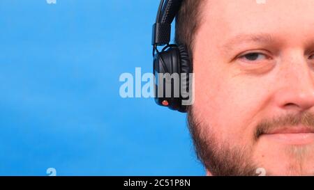 Bearded funny smiling cute fat man in a pink T-shirt listens to music and dances on a blue background, close-up, copy space. Stock Photo
