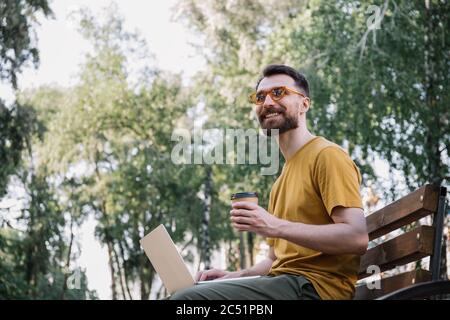 Young freelancer copywriter wearing stylish eyeglasses, working in park, typing. Man using laptop computer, holding cup of coffee, shopping online Stock Photo