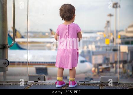 Cute Toddler baby in a pink dress looks at the planes at the airport. Waiting for a flight flight. back view Stock Photo