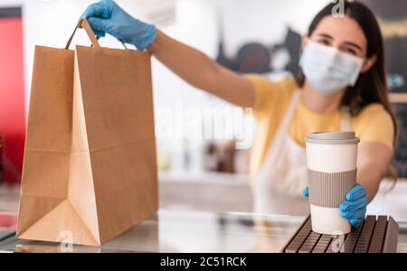 Young woman wearing face mask while serving takeaway breakfast and coffee inside cafeteria restaurant - Worker preparing delivery food inside bakery b
