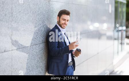 Business owner in formal suit drinking coffee and checking his phone near office building wall Stock Photo