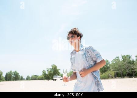 Taking champagne glasses. Seasonal feast at beach resort. Stylish boy celebrating, resting, having fun in sunny summer day. Looks happy and cheerful. Festive time, wellness, holiday, party. Stock Photo
