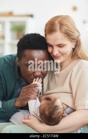 Vertical portrait of happy interracial family cuddling newborn baby, with African-American father kissing tiny hand Stock Photo