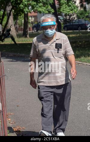 An older Asian American man, likely Chinese American, exercise walks in Kissena Park wearing both a surgical mask and a plastic face shield. Stock Photo