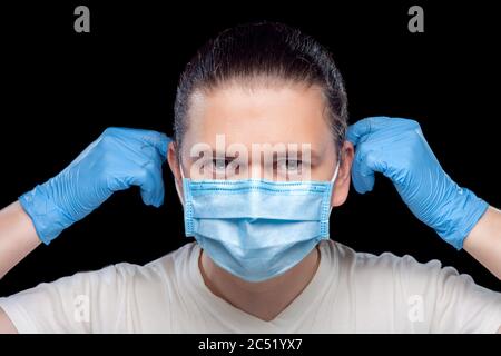 Portrait of a male doctor putting a medical mask on his face with hands in styry gloves, closeup of a medical worker isolated on black background. Stock Photo