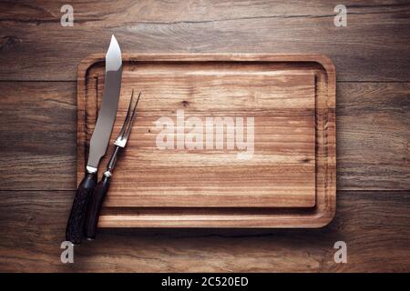 Empty vintage wooden cutting board with roast knife and fork on wooden table. Overhead view with copy space. Stock Photo