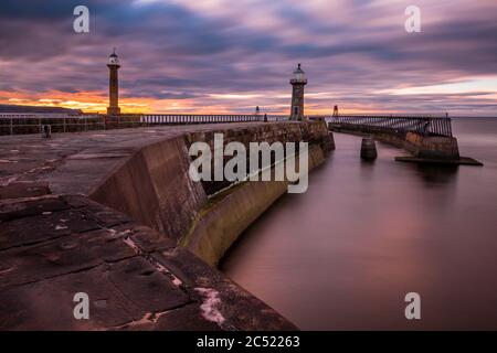 Dusk at Whitby harbour from the end of the pier. Warm sunset behind the lighthouses Stock Photo