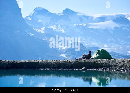 Man traveller enjoying scenery, sitting in chair near tent, admiring fantastic view of alpine mountains near lake with fresh clear water. Concept of travelling, hiking and camping in Alps. Stock Photo