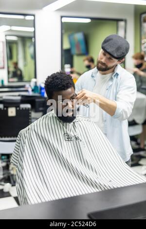 Happy young black man being trimmed with electric clipper machine in barbershop. Male beauty treatment concept. Smiling young African guy getting new Stock Photo